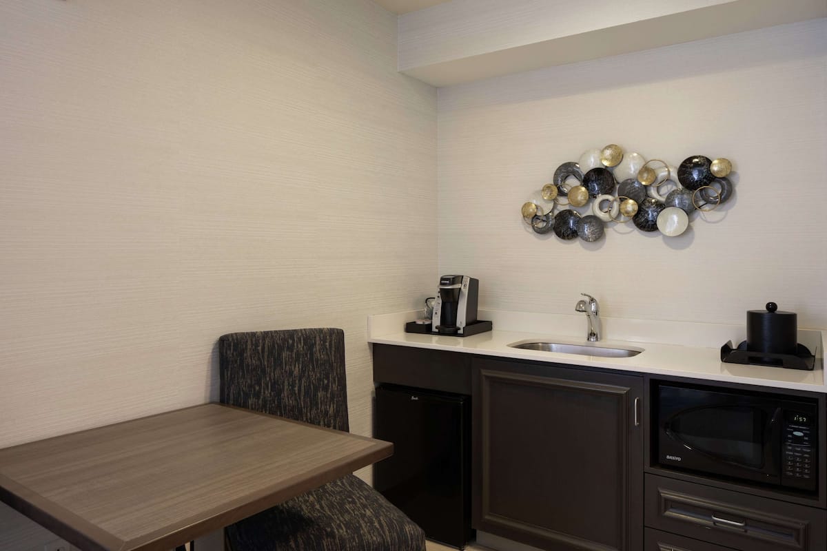 A compact kitchenette with a coffee machine, sink, microwave, and a seating area with a chair.