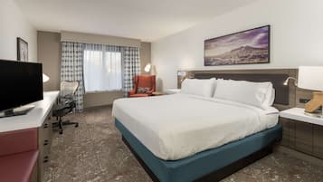 A hotel room with a bed, a desk with a TV, a chair, a lamp, and a painting on the wall.