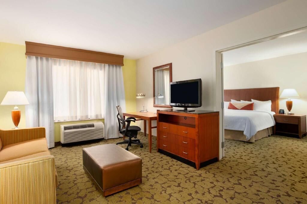 A hotel room with a bed, a desk with a computer, a television, a chair, an ottoman, and a lamp.