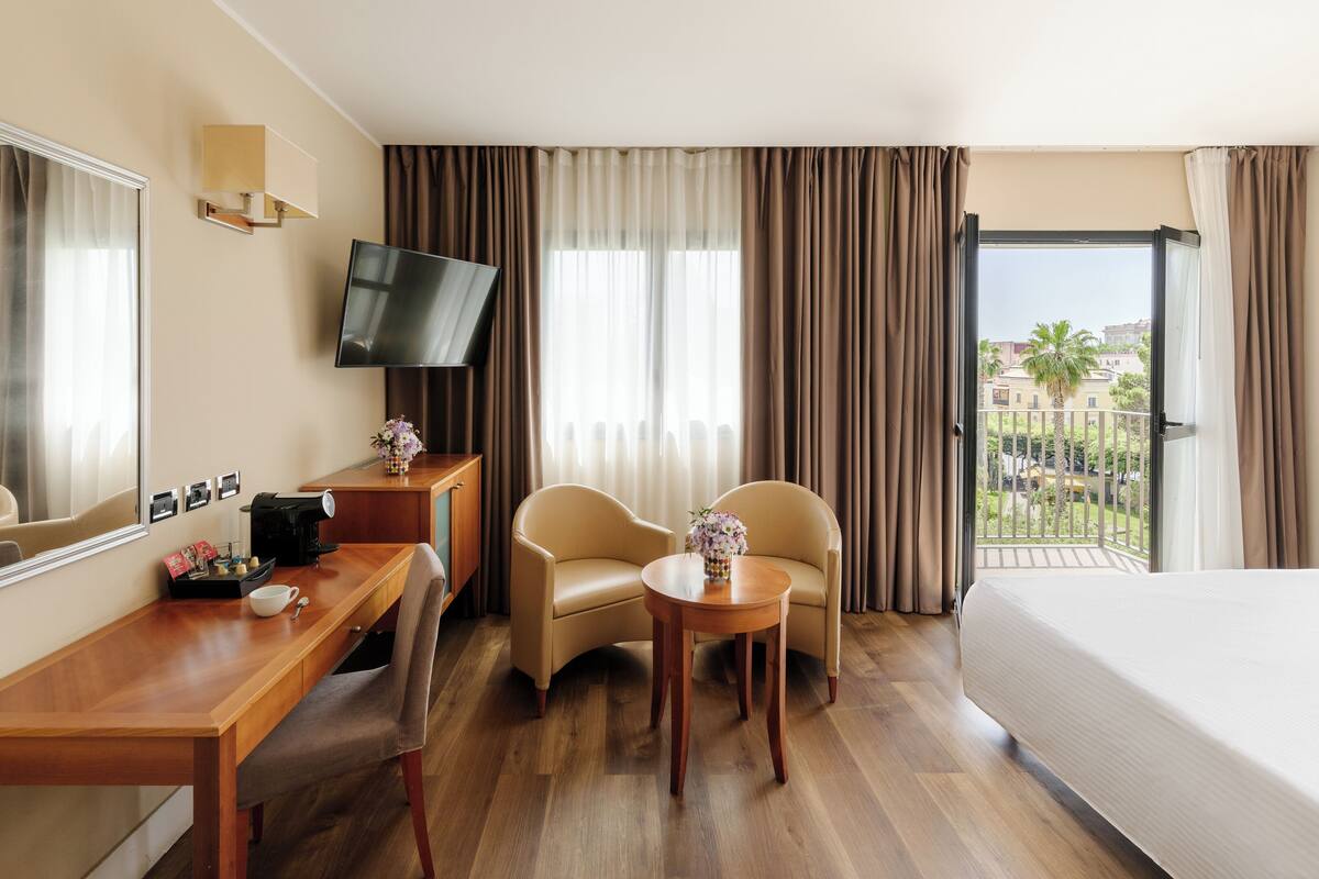 Une chambre d’hôtel comprenant un lit, un bureau avec une machine à café, deux chaises, une télévision et un balcon offrant une vue sur les immeubles et les palmiers.
