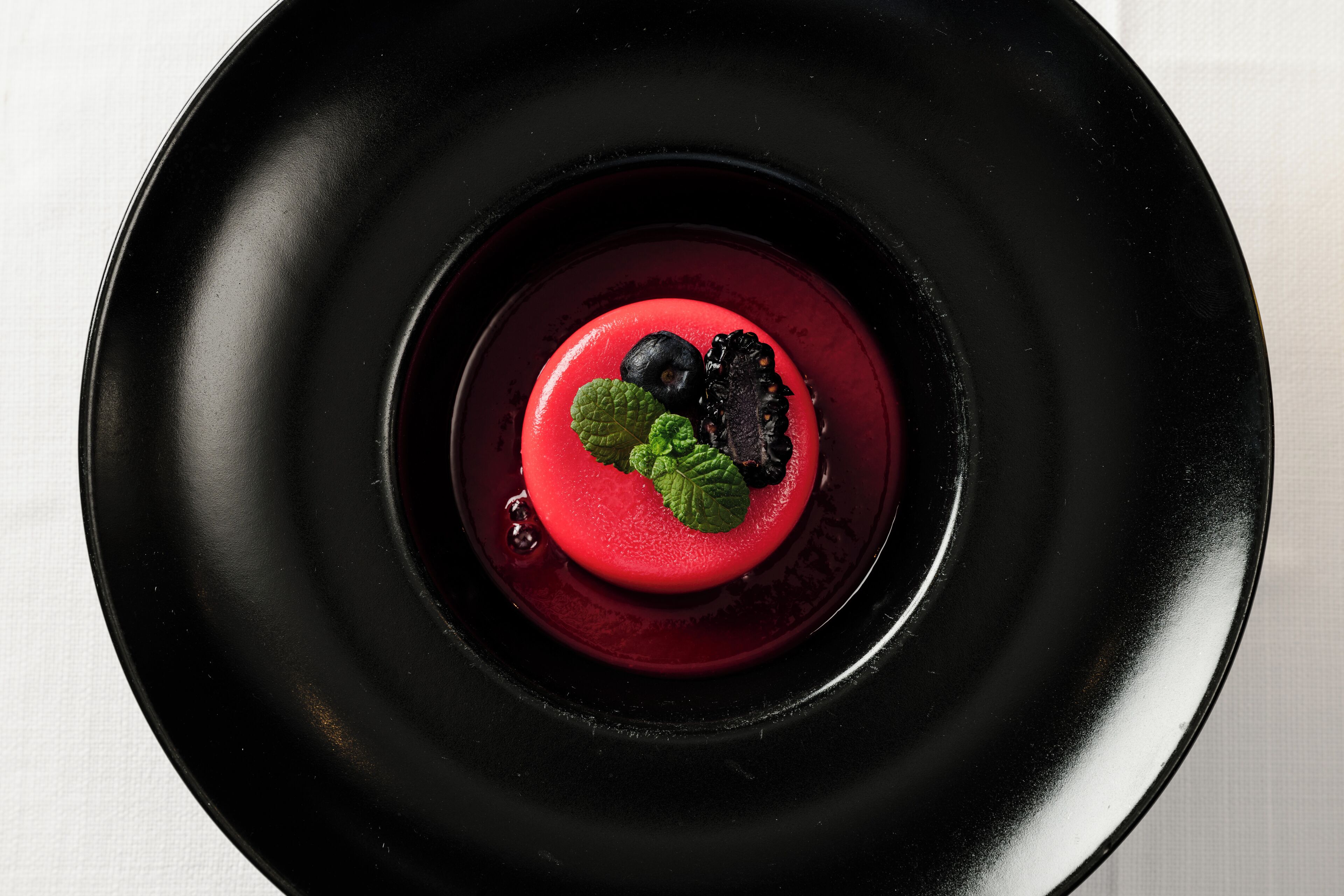 Un dessert su un piatto nero con base rossa e guarnito con foglie di menta e frutti di bosco.