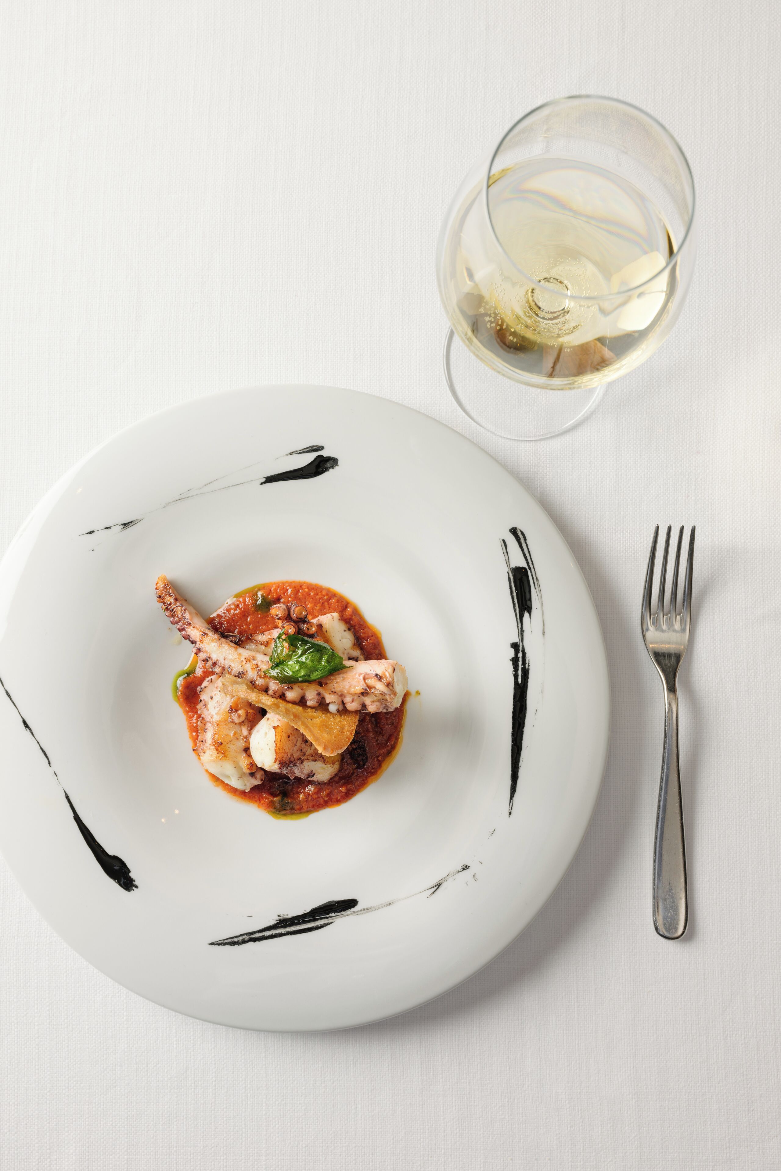 A white plate with a seafood dish, a fork, and a glass of white wine on a white tablecloth.