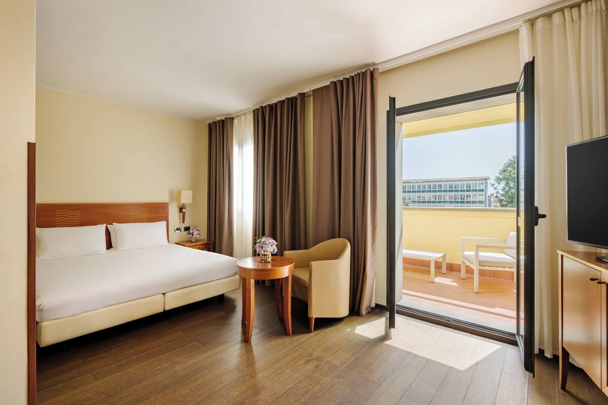 Une chambre d’hôtel avec un lit, une petite table sur laquelle est posé un vase, une chaise, un téléviseur et un balcon offrant une vue sur les bâtiments.