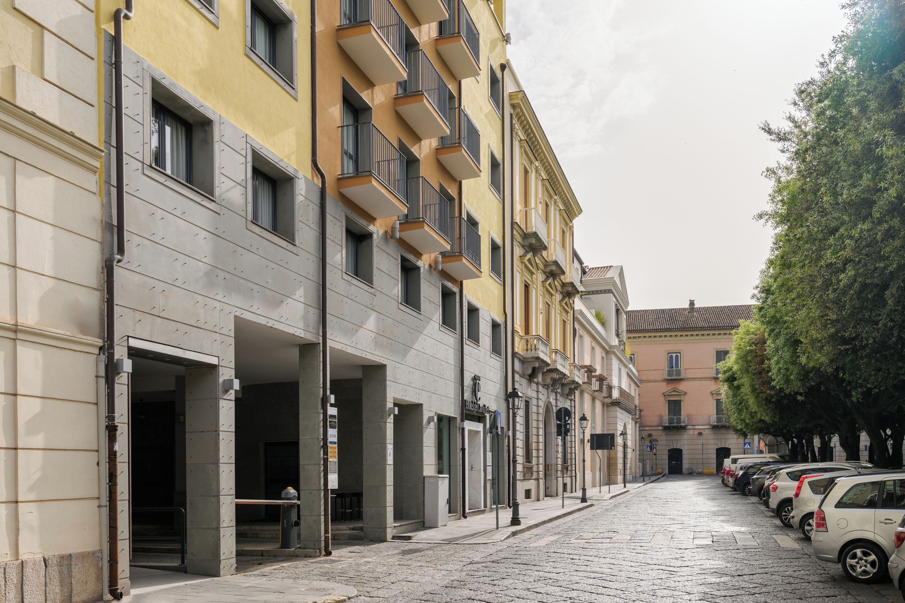 A row of parked cars along a cobblestone street with buildings featuring balconies and a clear sky.