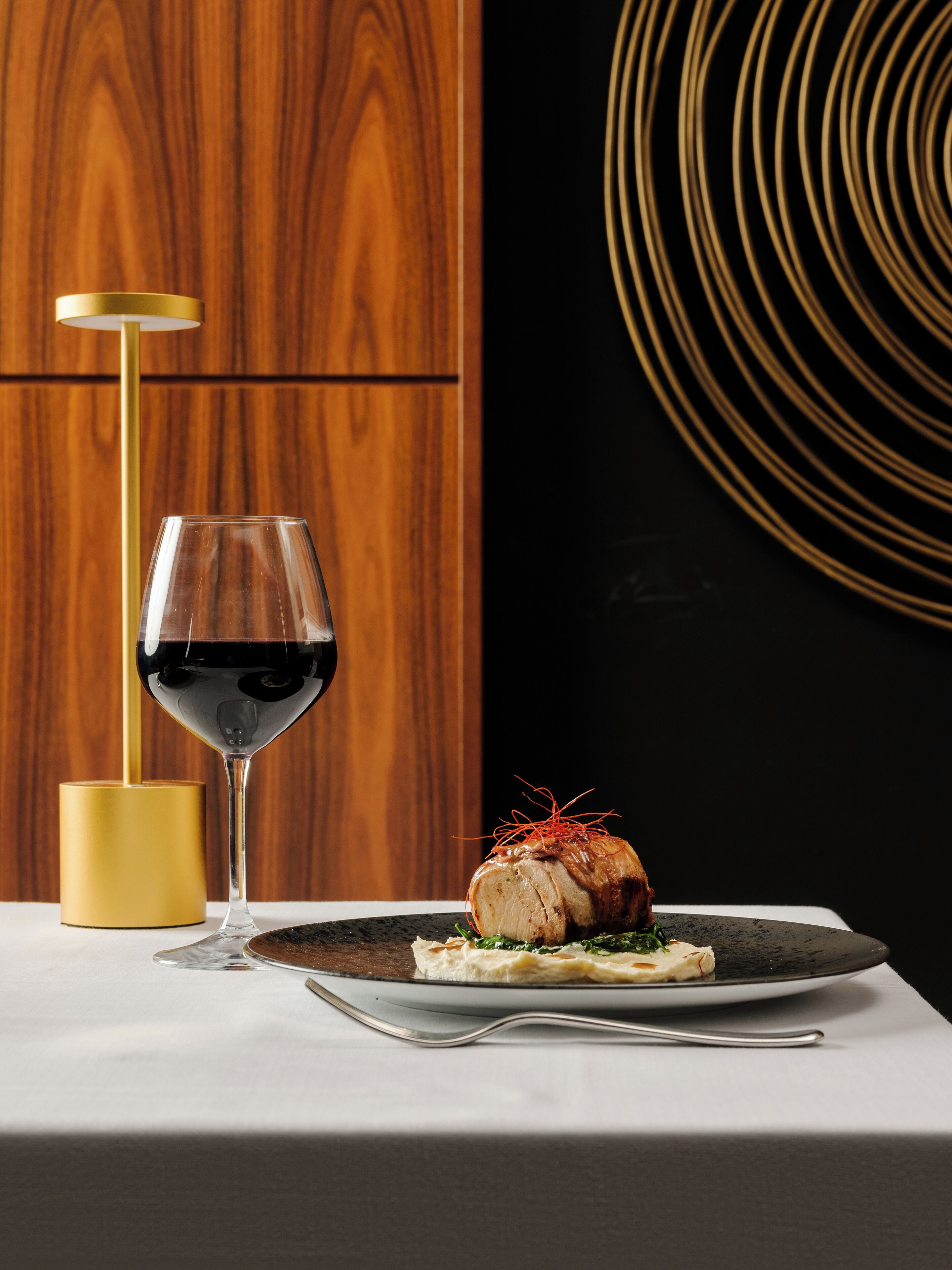 Una mesa de comedor con un plato de comida, una copa de vino y una lámpara dorada.