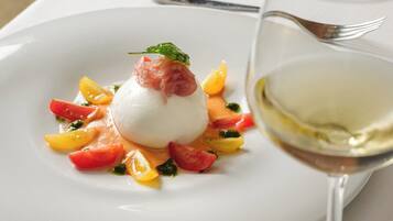 Um prato branco com um alimento redondo e branco, coberto por uma folha verde, rodeado por legumes coloridos e um copo de vinho branco.