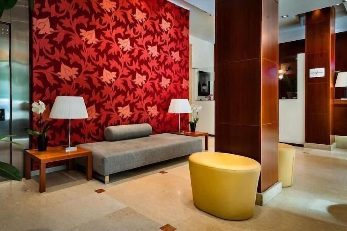 Un hall d’entrée avec un mur orné d’un motif rouge, un canapé gris, deux tables d’appoint avec des lampes, un pouf jaune et une grande colonne en bois.