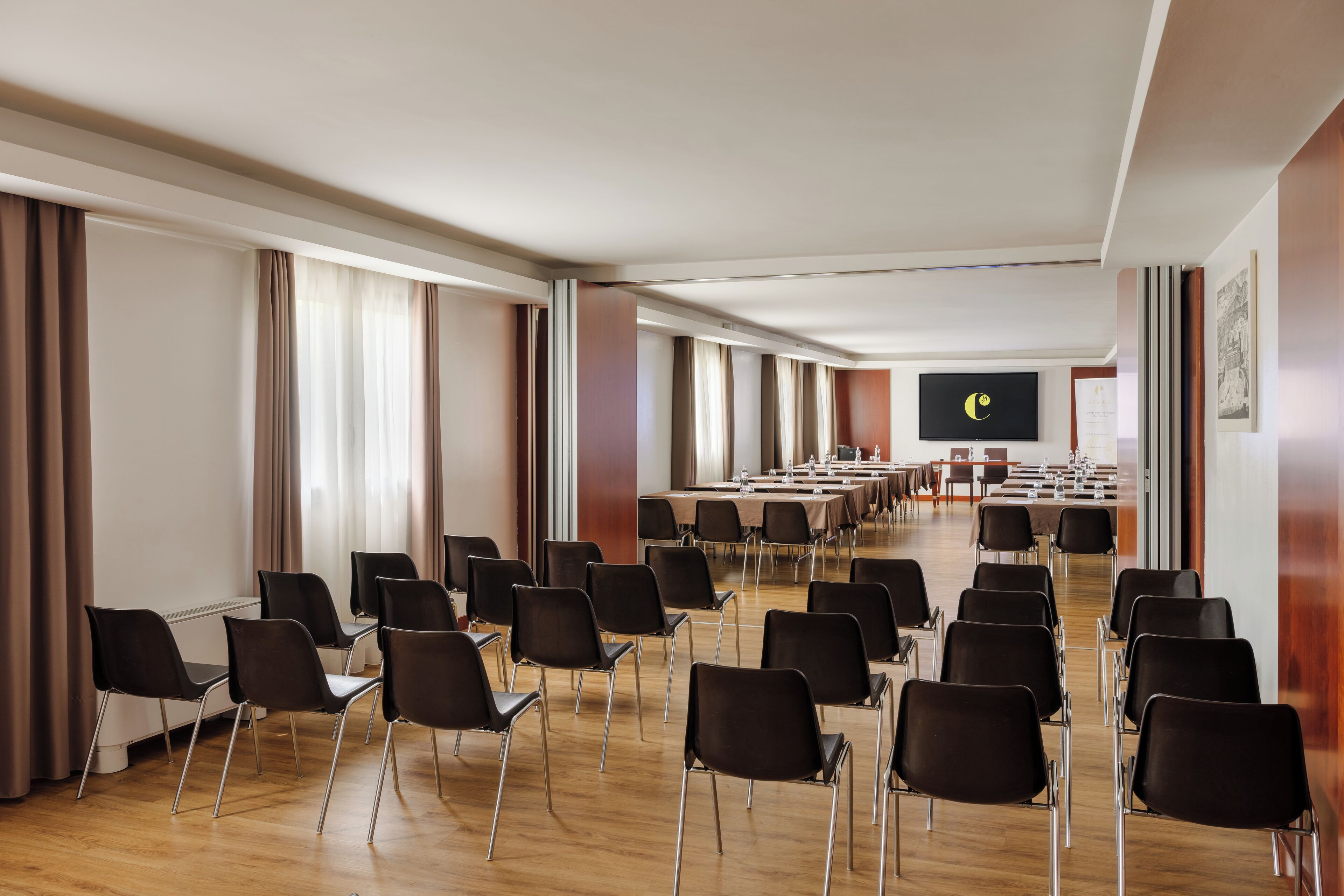 Un salón de conferencias con filas de sillas, una mesa larga, una pantalla de proyector y un amplio ventanal con cortinas.