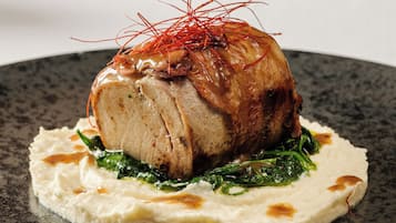 Un piatto impiattato con carne arrosto al centro, guarnito con fili rossi, su un letto di verdure a foglia verde e una base cremosa, su un piatto scuro e variegato.