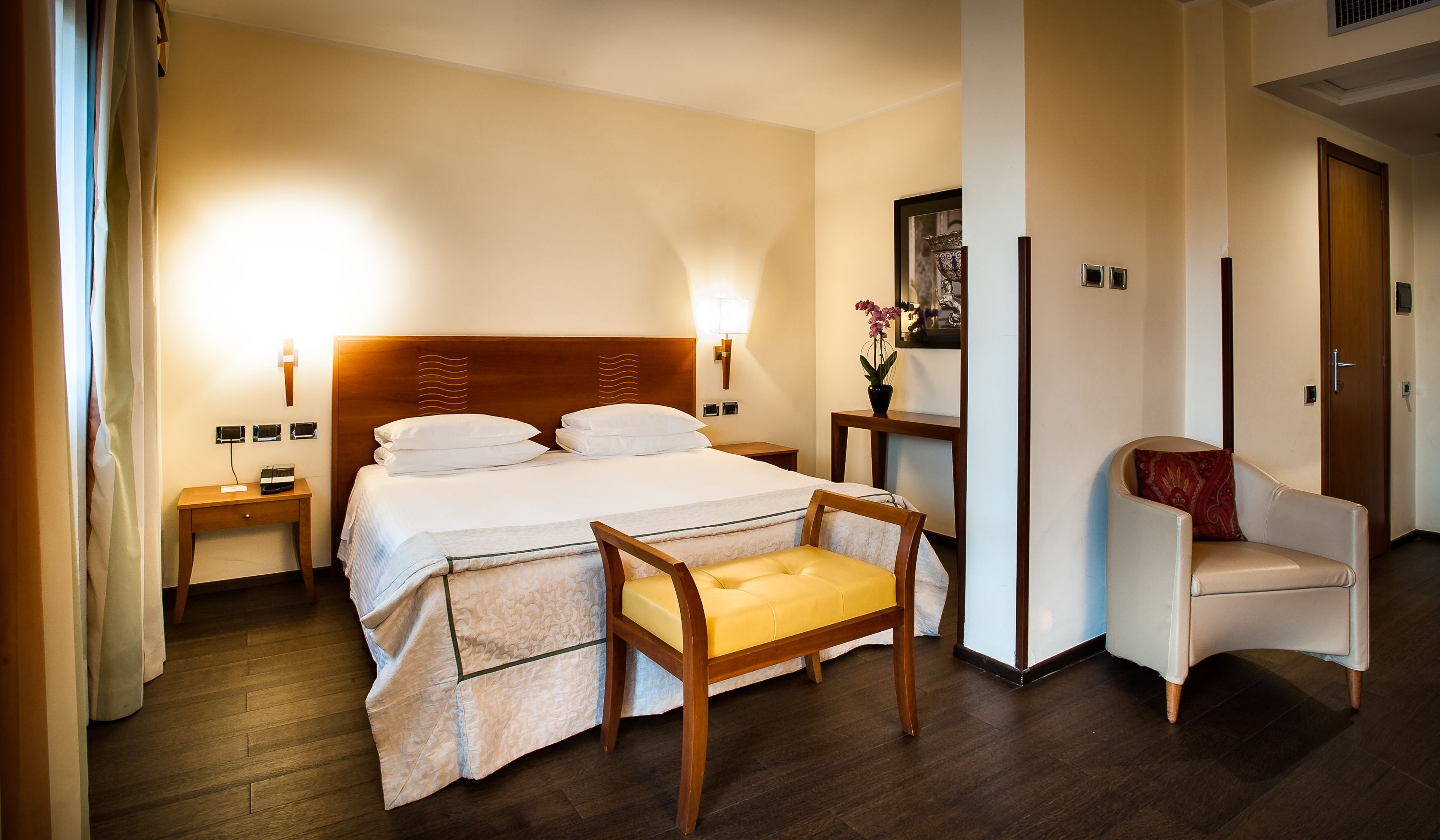 Una habitación de hotel con una cama, mesitas de noche, una silla y una pequeña mesa con un florero.
