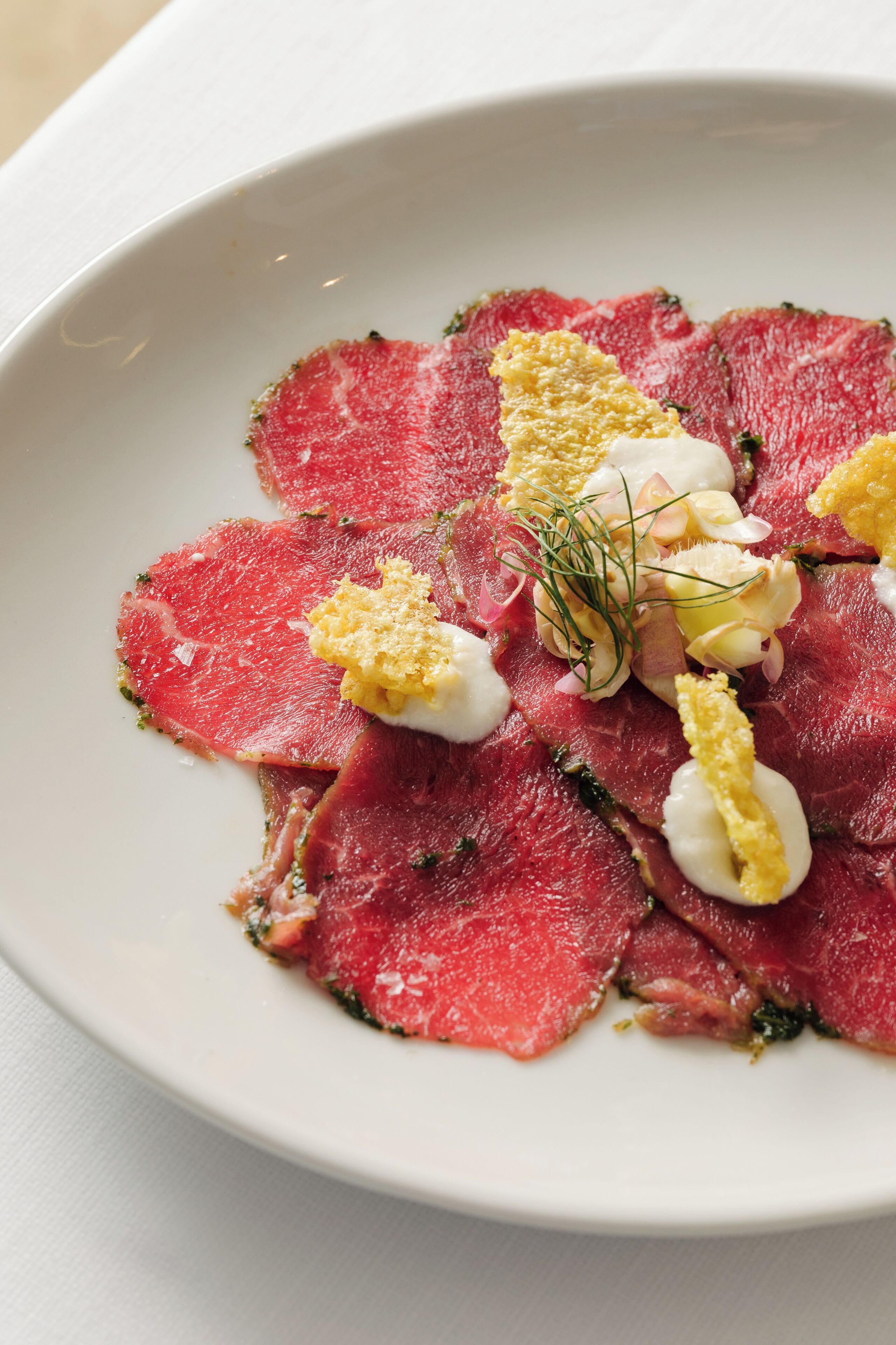 Um prato branco com carne vermelha fatiada finamente, decorado com ervas e pequenos itens amarelos.