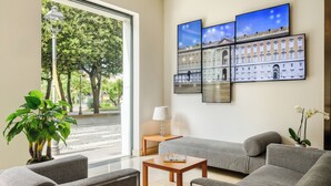 Ein modernes Hotelzimmer mit großem Fenster, einer grauen Couch, einem Holztisch und einem an der Wand montierten Fernseher, der eine Stadtansicht zeigt.