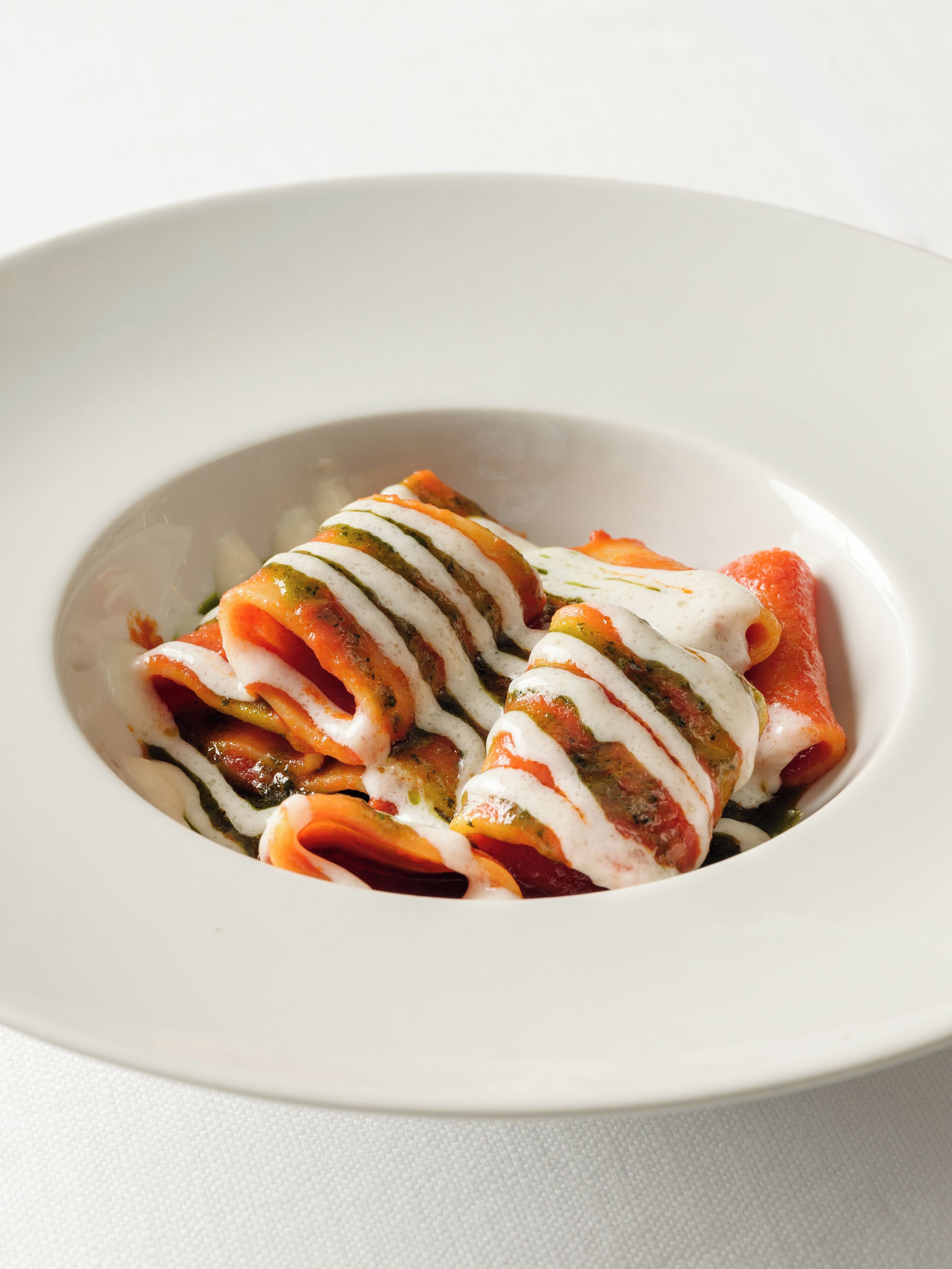 A white plate with a dish of rolled pasta topped with a white sauce and green herbs.
