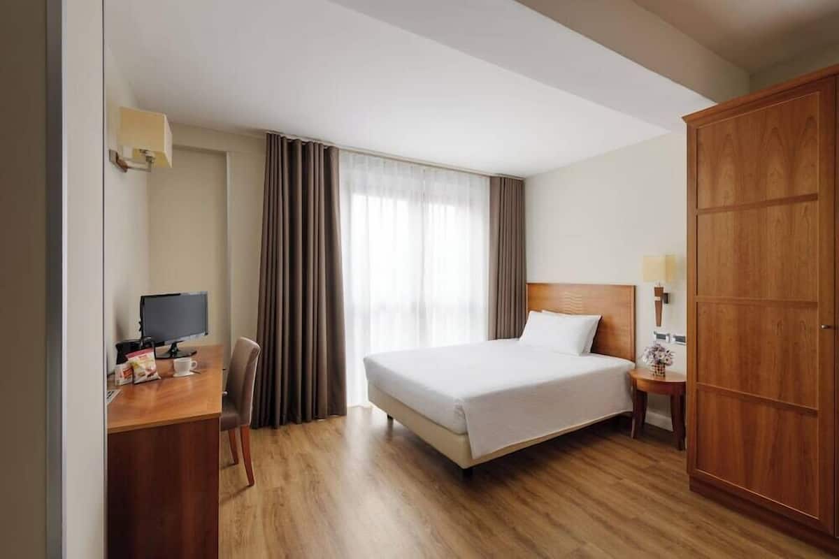 Une chambre d’hôtel comprenant un lit, un bureau avec un ordinateur, une chaise, une armoire et une fenêtre avec des rideaux.