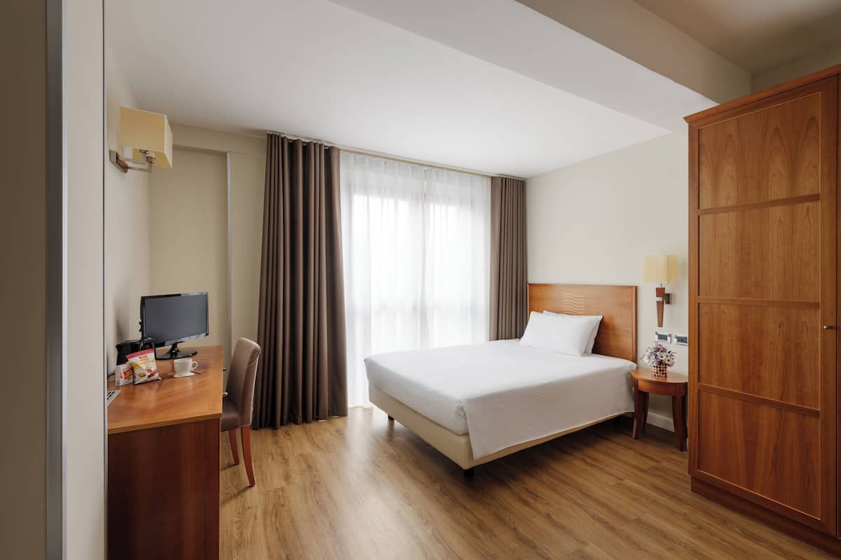 Une chambre d’hôtel avec un lit, un bureau avec un ordinateur, une chaise, une armoire en bois et une fenêtre avec des rideaux.