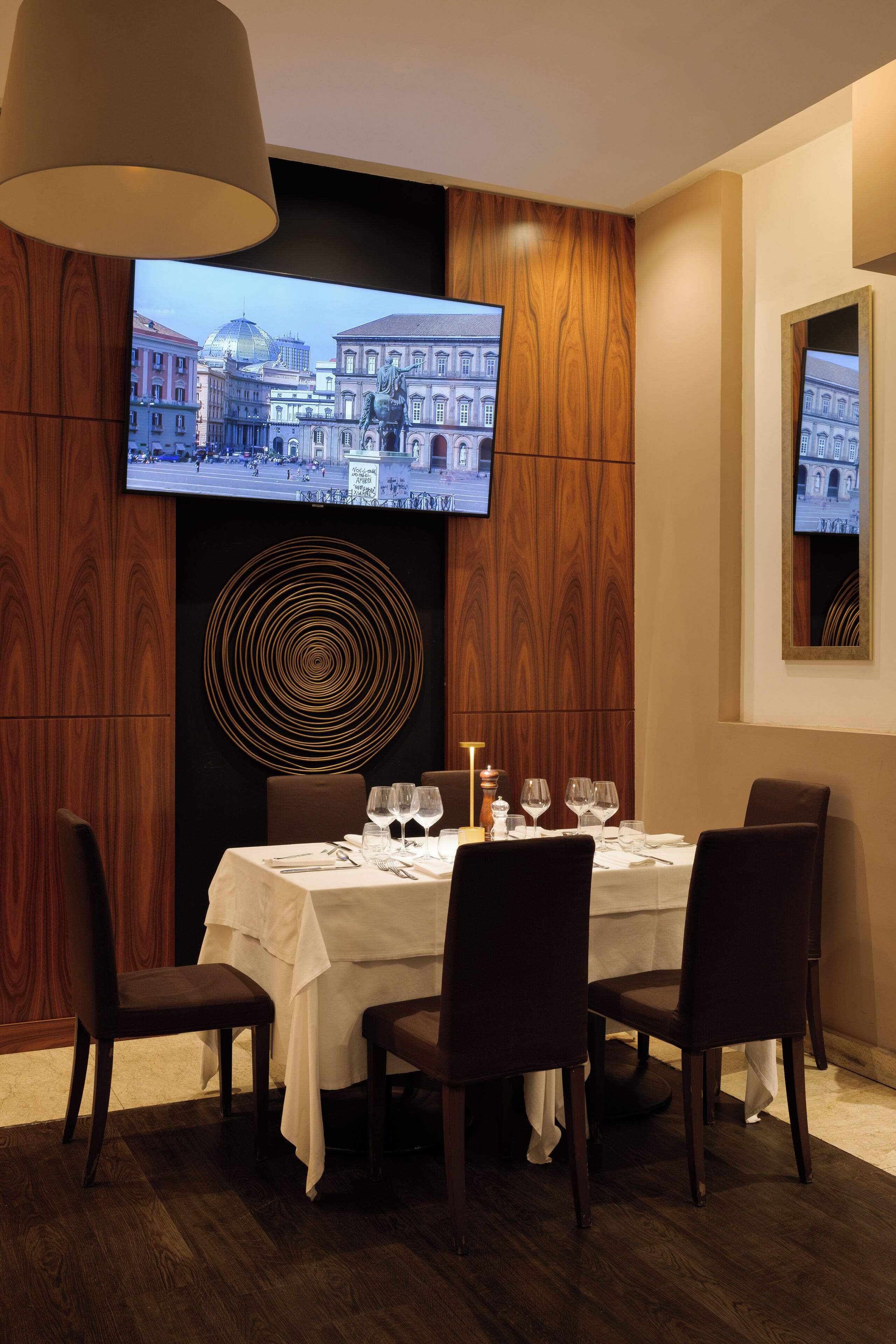 Un área de comedor con una mesa preparada para cuatro personas, un televisor de gran tamaño montado en la pared mostrando un paisaje urbano, y un adorno circular en la pared.
