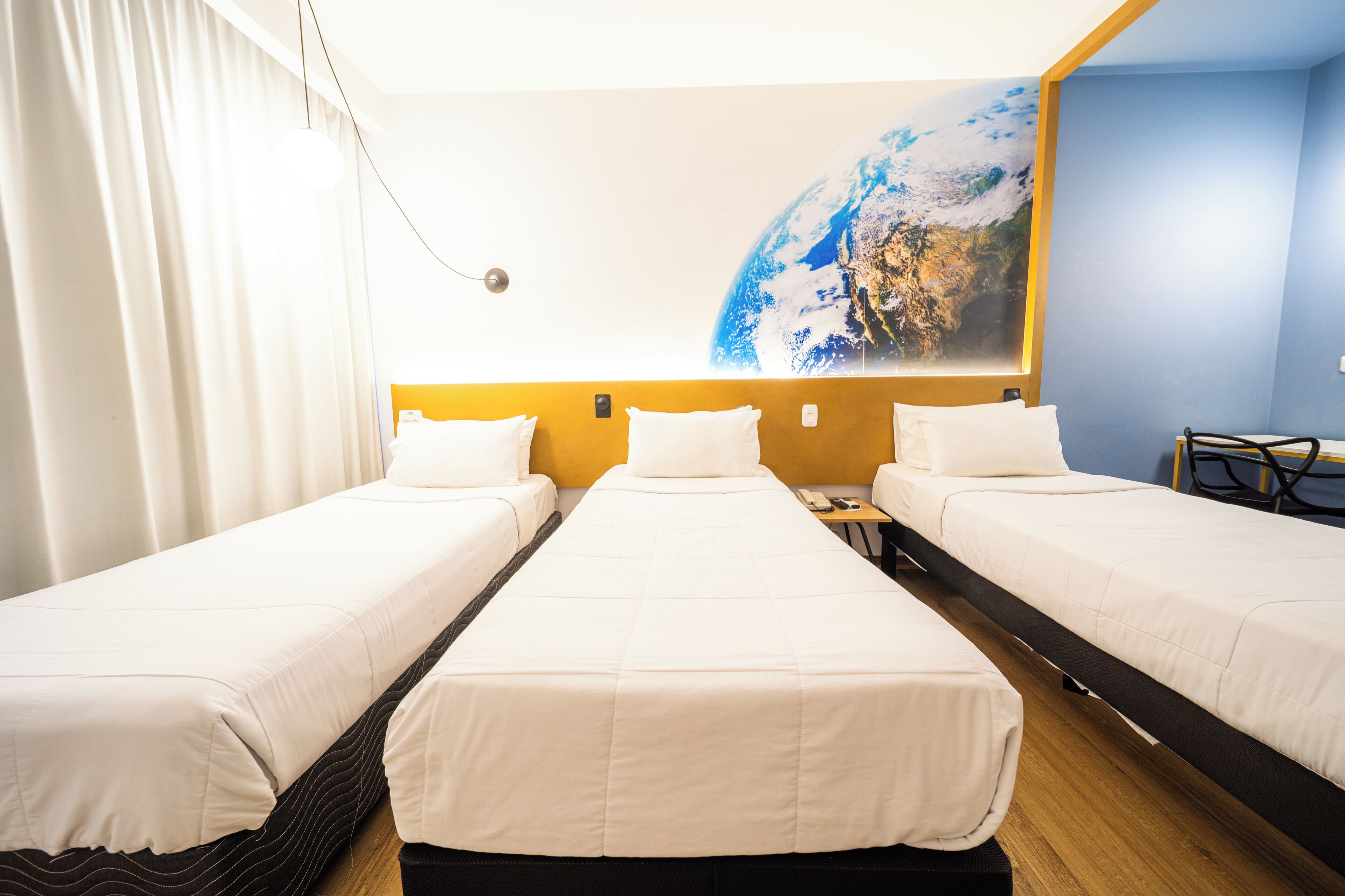 Habitación de hotel con tres camas individuales, un ventanal con vistas de la Tierra y un escritorio con una silla.