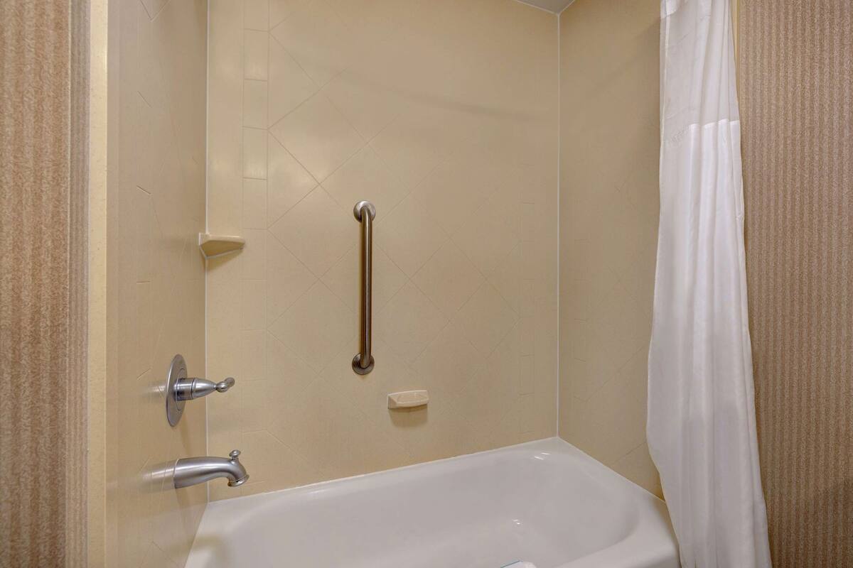 A bathroom with a bathtub, a handrail, a shower curtain, and a toilet.