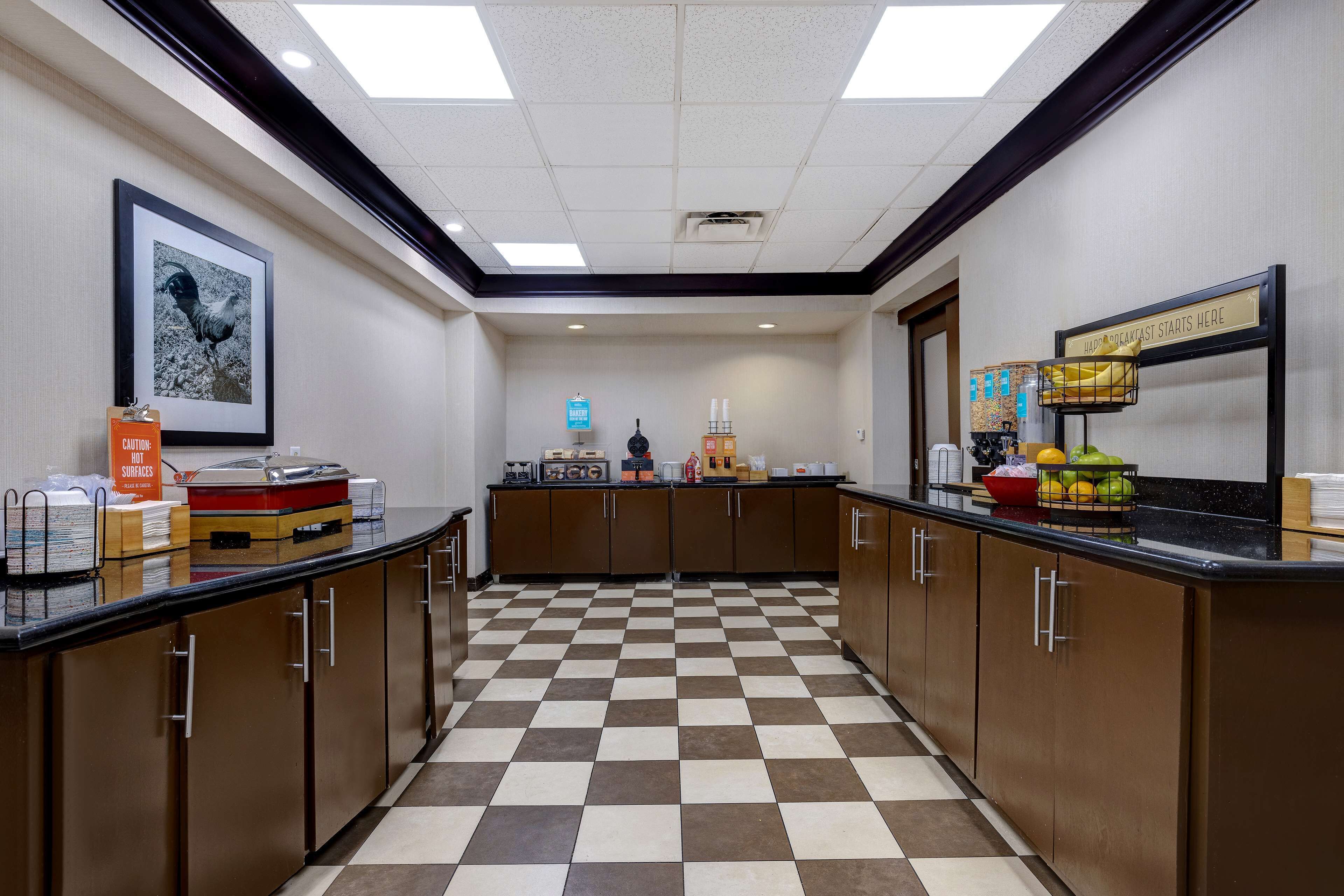 A hotel kitchen with checkered flooring, wooden cabinets, and a sign indicating breakfast is served.