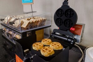 A waffle iron with four waffles in the process of cooking, a display case with various pastries, and a digital display indicating the temperature.