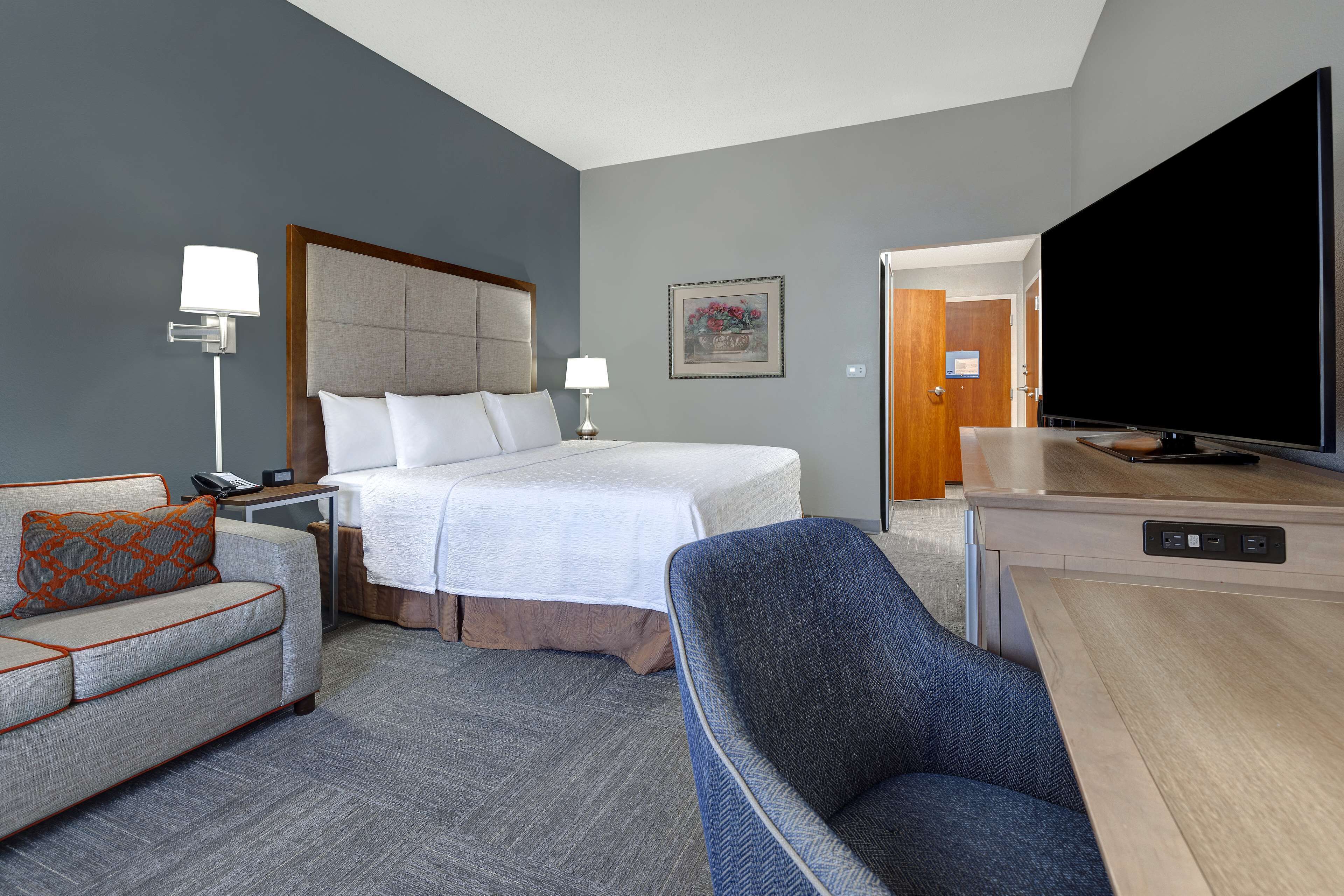 Habitación de hotel con cama, sofá, silla, televisor y escritorio con lámpara.
