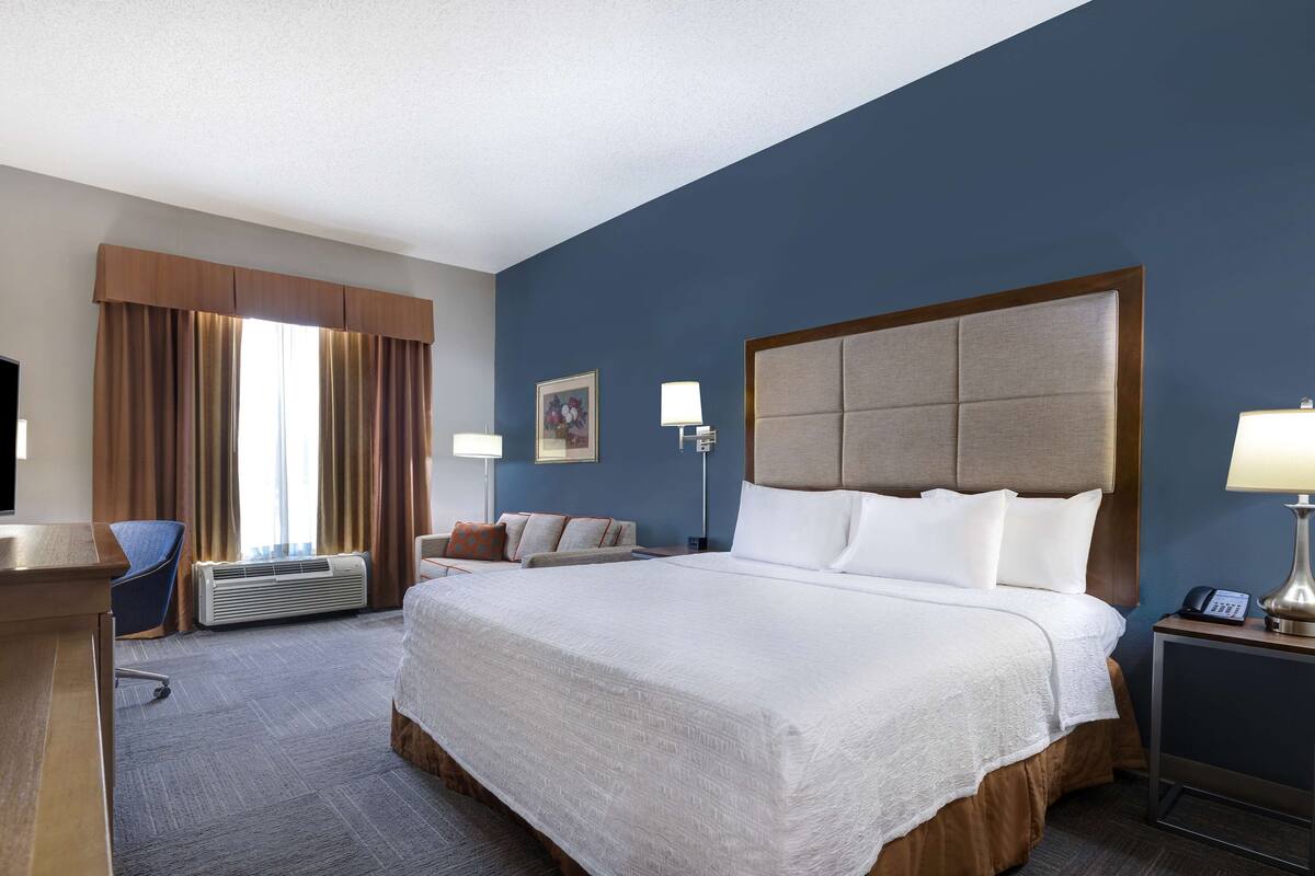 A hotel room with a large bed, a desk, a chair, a bedside table with a lamp, and a wall-mounted TV.