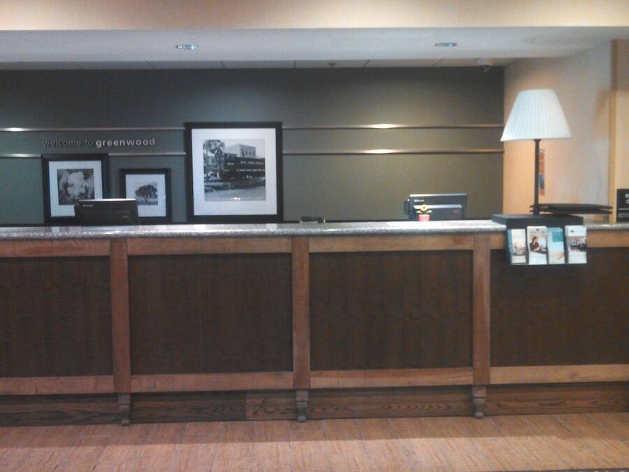 A reception desk with framed pictures, a "Welcome to Greenwood" sign, a "Manager On Duty" sign, and a lamp.