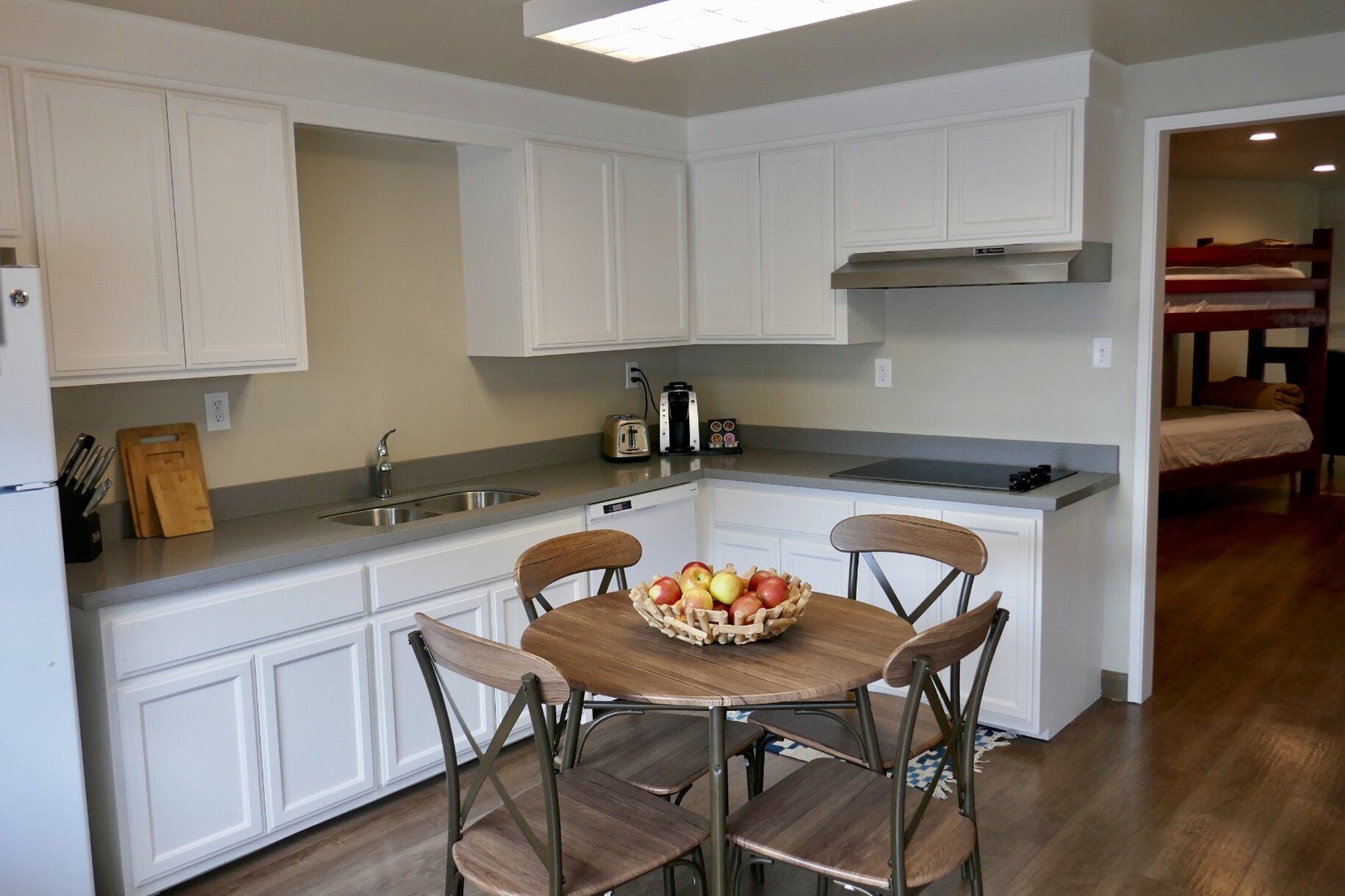 A compact kitchen with white cabinetry, a built-in stove, and a dining area with four chairs and a fruit bowl.