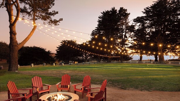Un foyer est entouré de chaises Adirondack rouges, le tout installé dans un espace extérieur, avec des guirlandes lumineuses suspendues au-dessus.