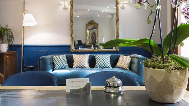 A hotel reception area with a blue sofa, a wooden cabinet, a mirror, and a potted orchid. - Hotel Mogador Opera - Paris (Paris)