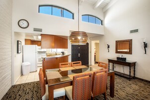 A spacious dining area with a glass table and patterned chairs, a kitchen in the background, and a large window providing natural light.