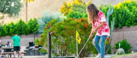 A person playing mini-golf in an outdoor setting with a flagstick, a flag, and a golf ball.