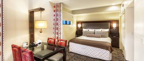 A hotel room with a bed, a desk with chairs, a lamp, and a patterned carpet.