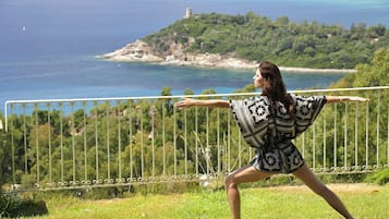 Una persona realizando una postura de yoga en una colina cubierta de hierba, con una vista panorámica de la costa y una torre al fondo.
