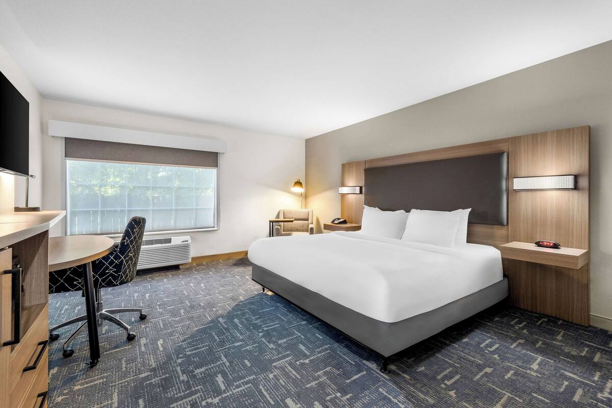 A hotel room with a large bed, a desk, a chair, a nightstand, a lamp, and a window with blinds.