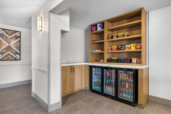 A kitchenette with wooden cabinets, a built-in shelf with snacks, and two mini-fridges stocked with beverages.