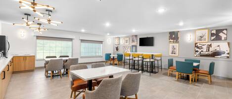 A modern dining area with a central table and surrounding chairs, a kitchen area with wooden cabinets, and a television mounted on the wall.