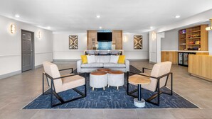 Un hall d’entrée moderne avec un canapé gris, deux fauteuils, une table basse et un coin cuisine doté de meubles en bois et d’un réfrigérateur.