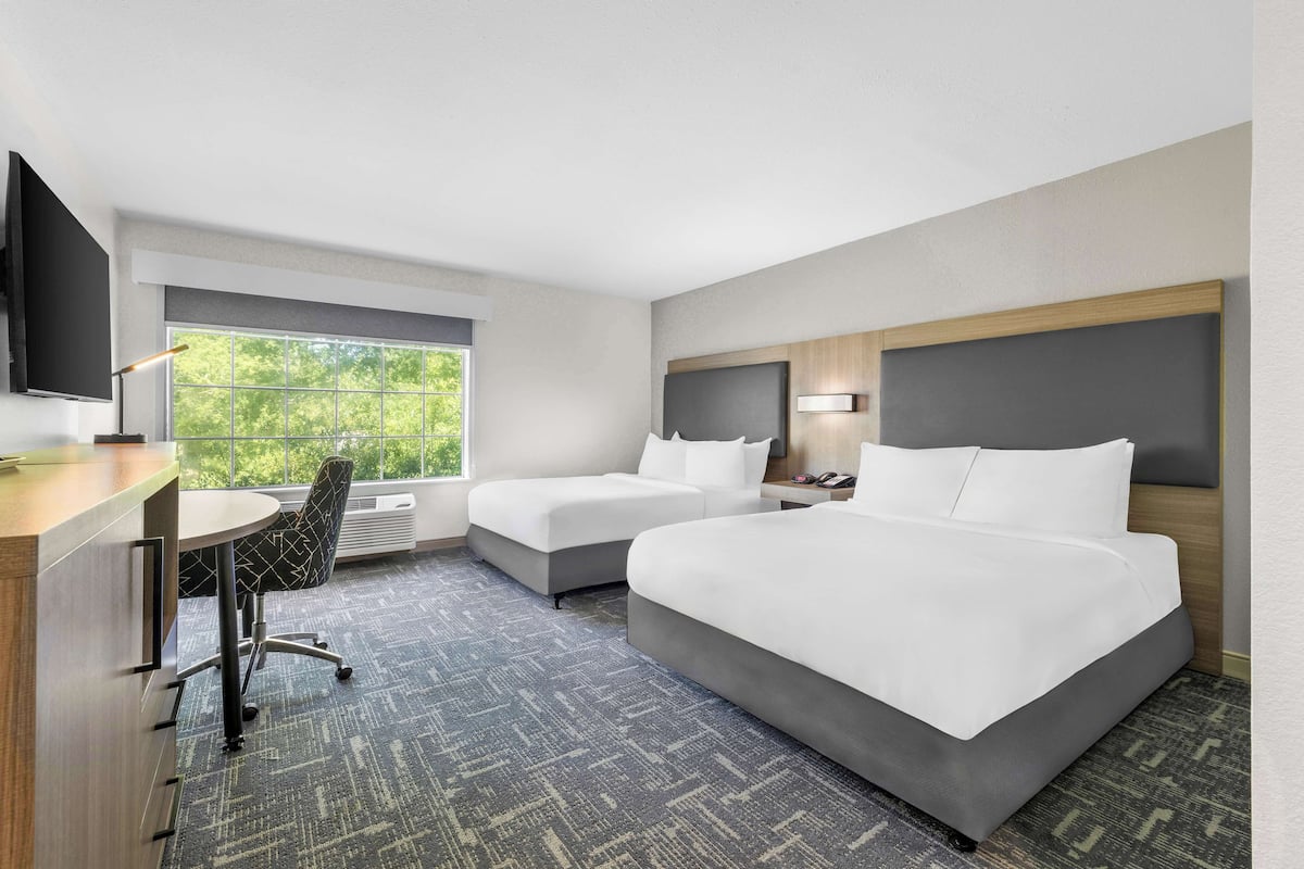A hotel room with two beds, a desk, a chair, a TV, and a large window with a view of greenery.