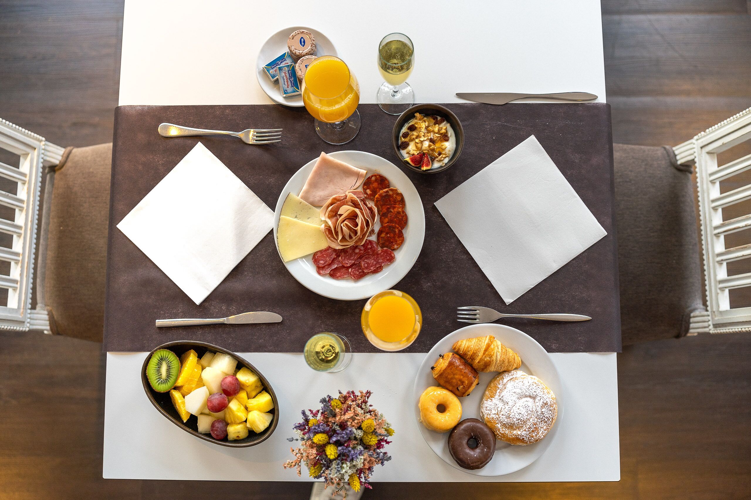A breakfast table with plates of food, a fruit bowl, a vase of flowers, and a glass of orange juice.