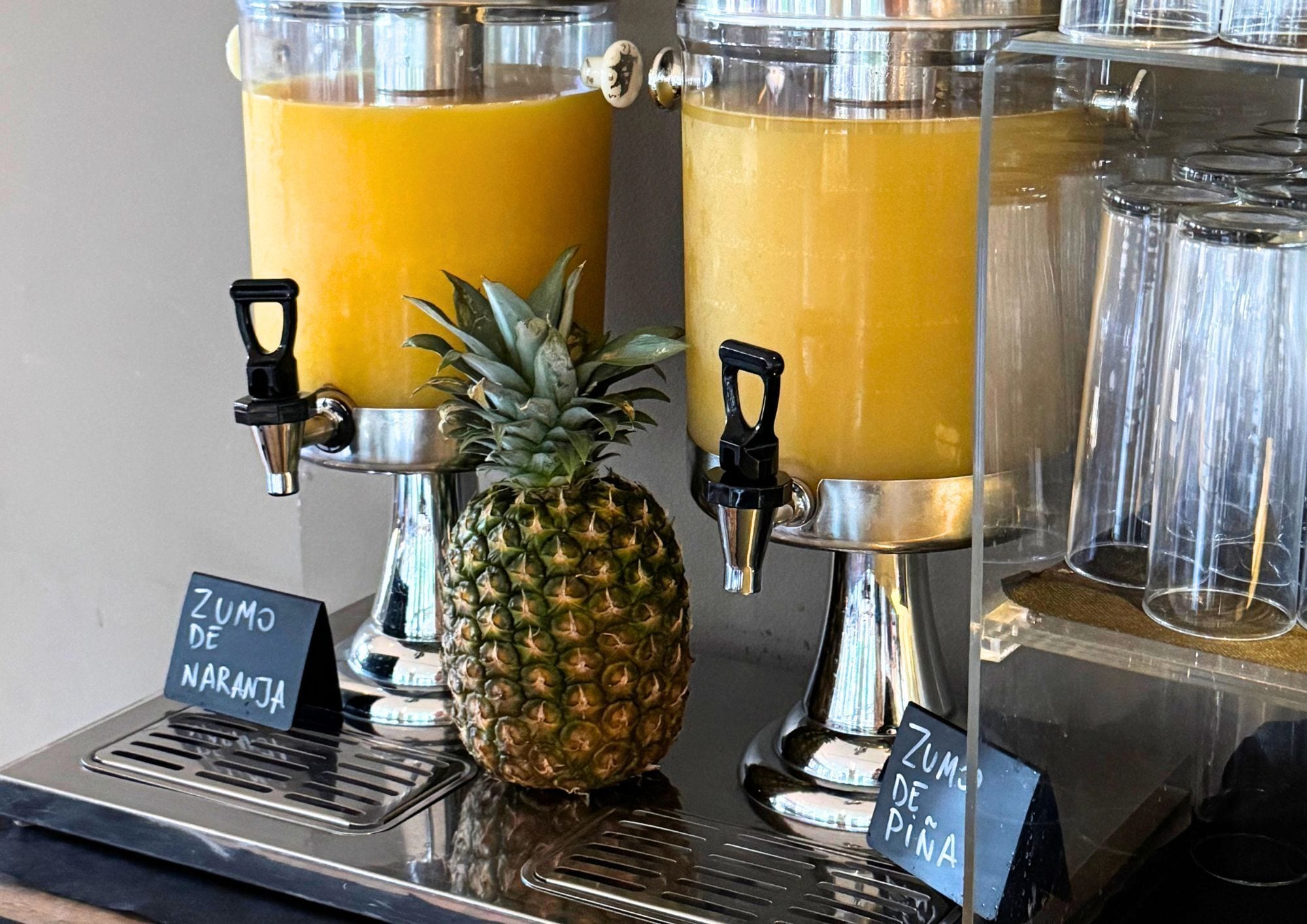 Two beverage dispensers with labels 'Zumo de Naranja' and 'Zumo de Piña' and a pineapple between them.