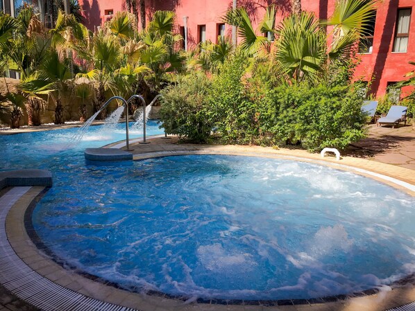 Una piscina con una cascada, rodeada de abundante vegetación y un edificio rojo al fondo.