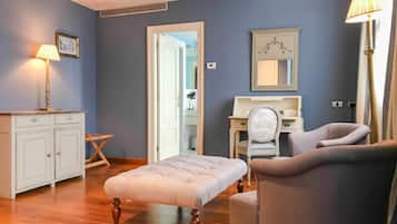 A room with a wooden floor, a white bench, a grey armchair, a white dresser, a vanity with a mirror, and a lamp.