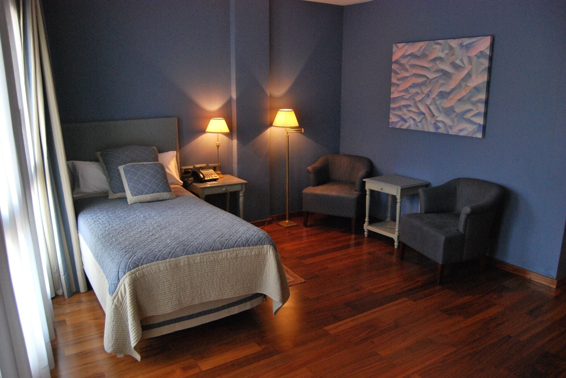 Una camera d'albergo con un letto, due poltrone, un comodino con una lampada e un quadro appeso al muro.