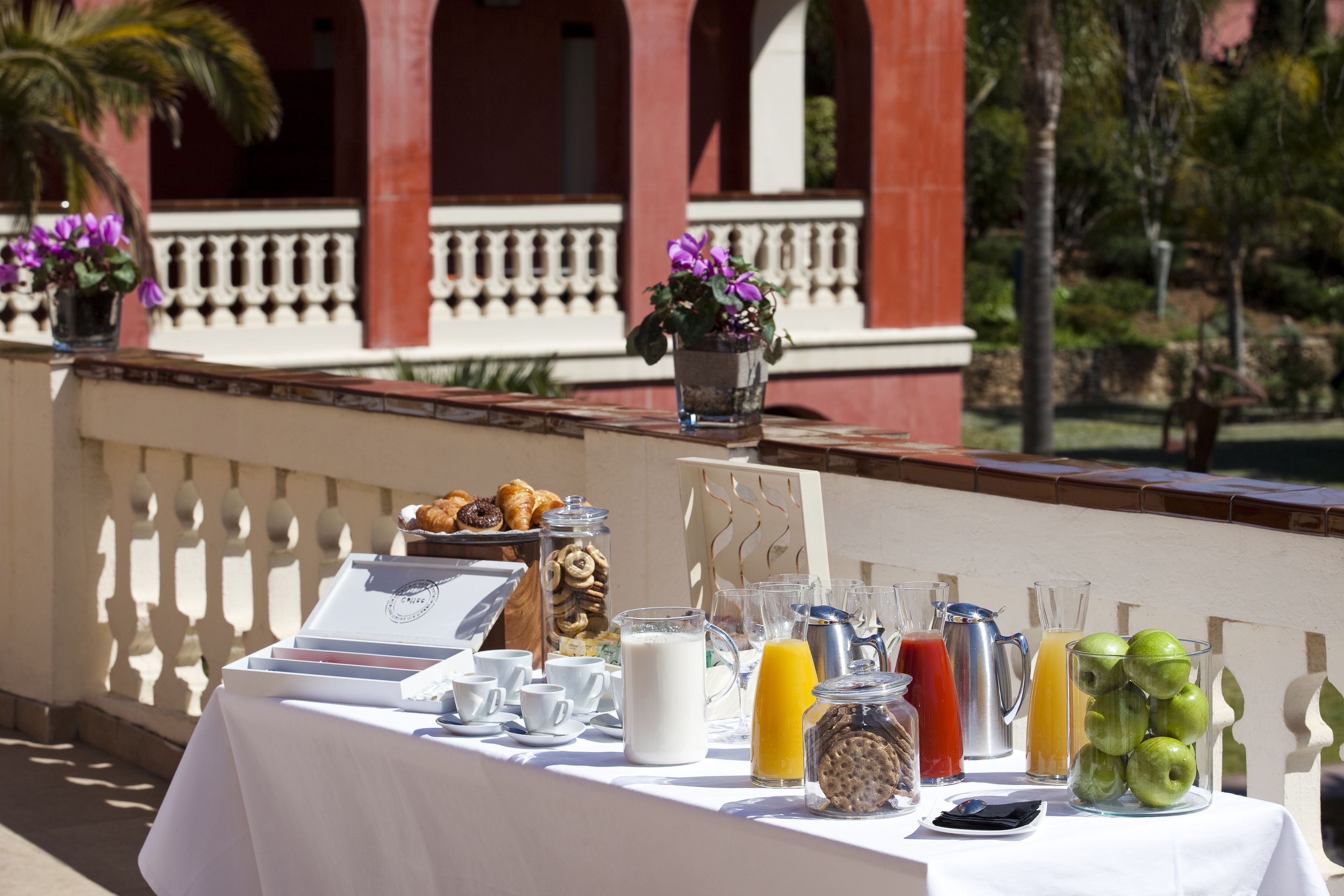 Une table de buffet proposant divers aliments et boissons pour le petit-déjeuner, placée devant un bâtiment doté d’un balcon et bordé de palmiers.