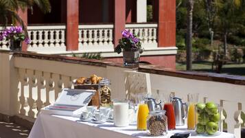 Une table de buffet proposant divers aliments et boissons pour le petit-déjeuner, placée devant un bâtiment doté d’un balcon et bordé de palmiers.