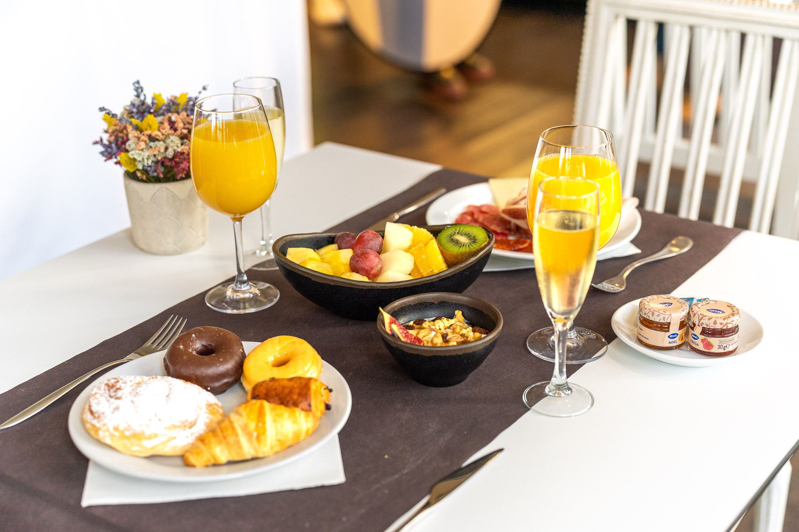 A breakfast table with croissants, a bowl of fruit, two glasses of orange juice, and a plate of jam.