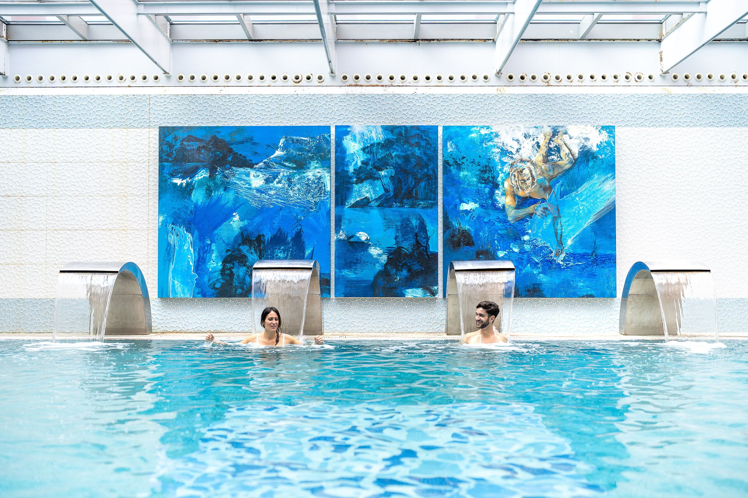 A swimming pool with two people in the water, surrounded by large blue murals on the wall.