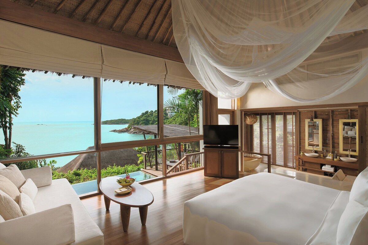 A spacious room with a large window offering a view of the sea, a comfortable sofa, a TV, and a wooden vanity with a mirror.