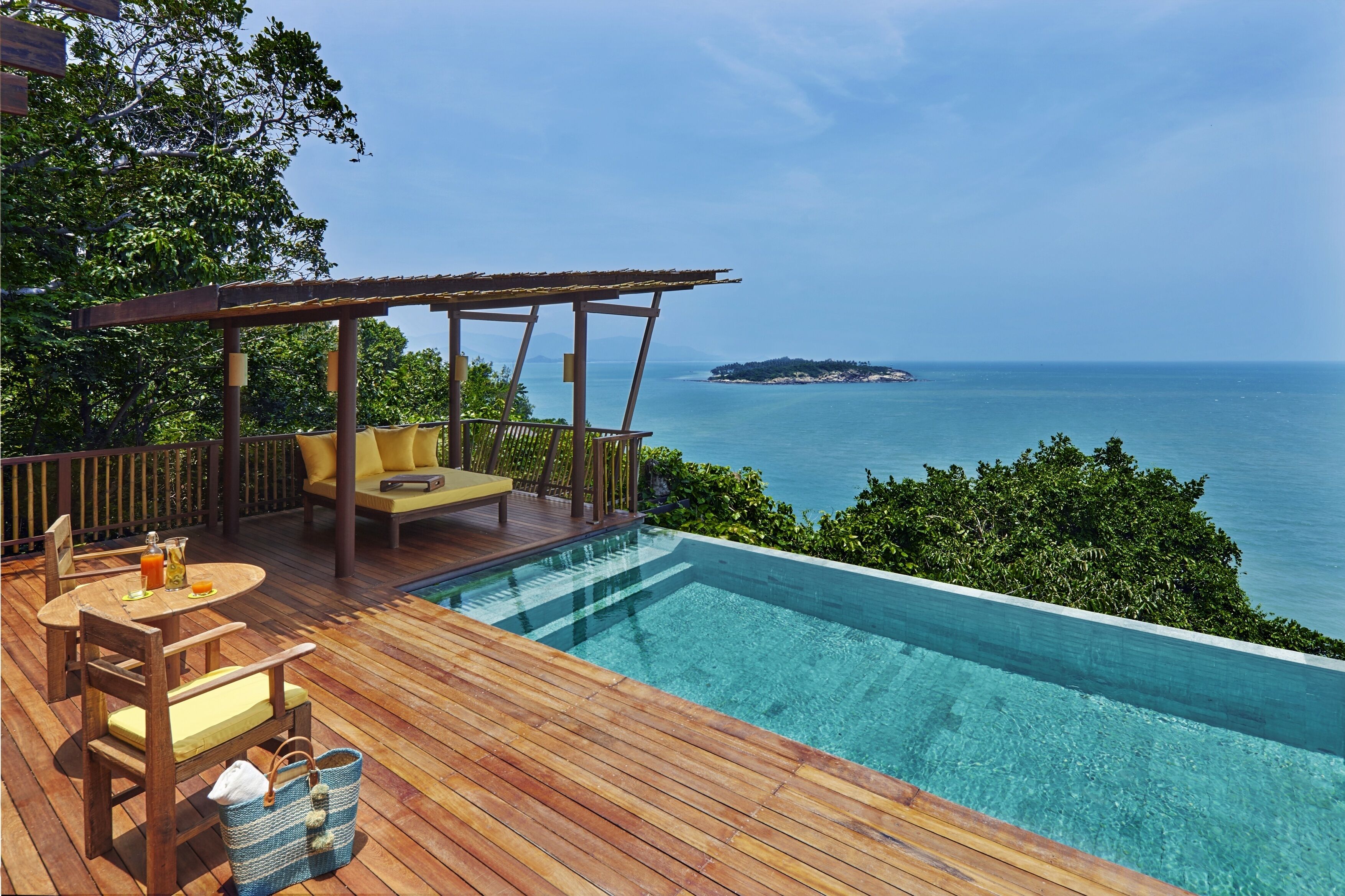 Terraza de madera con piscina con vistas al mar, una pérgola y un área de descanso con mesa y sillas.